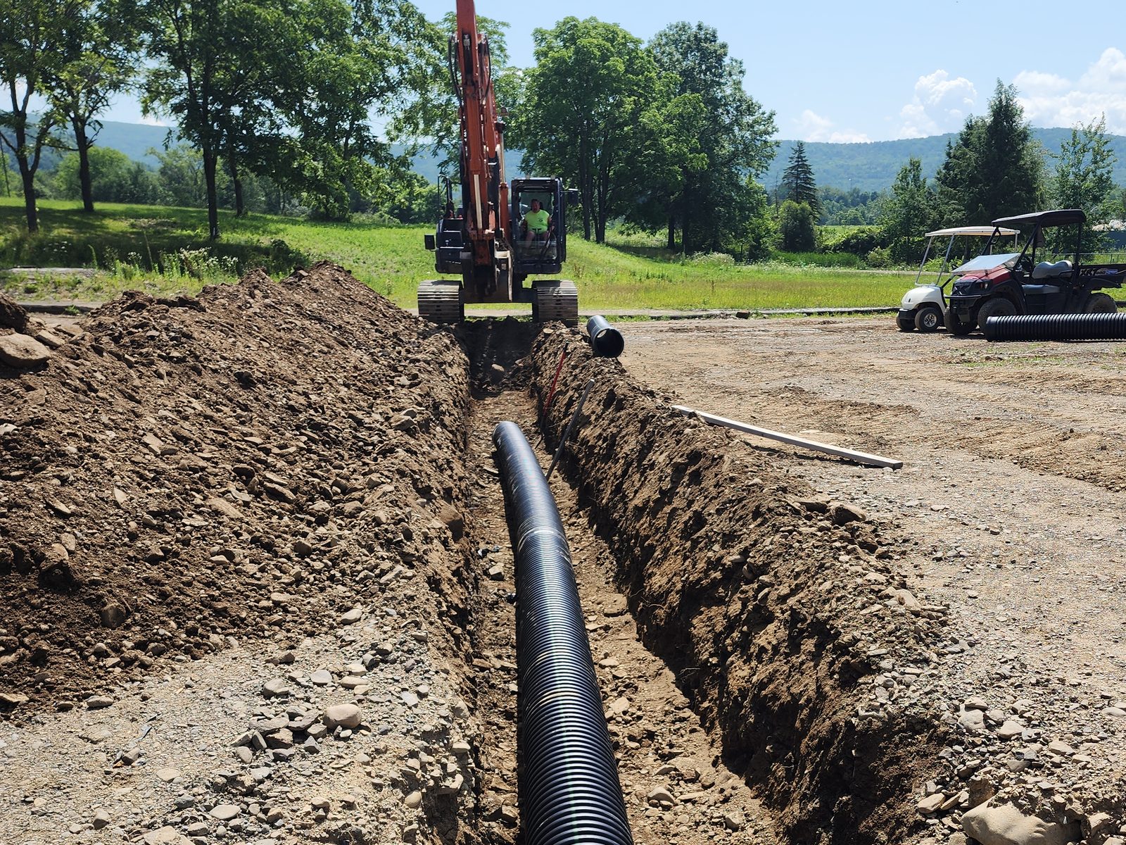 Large black drainage pipe laid in a long trench, excavator at the far end and wooded hills behind