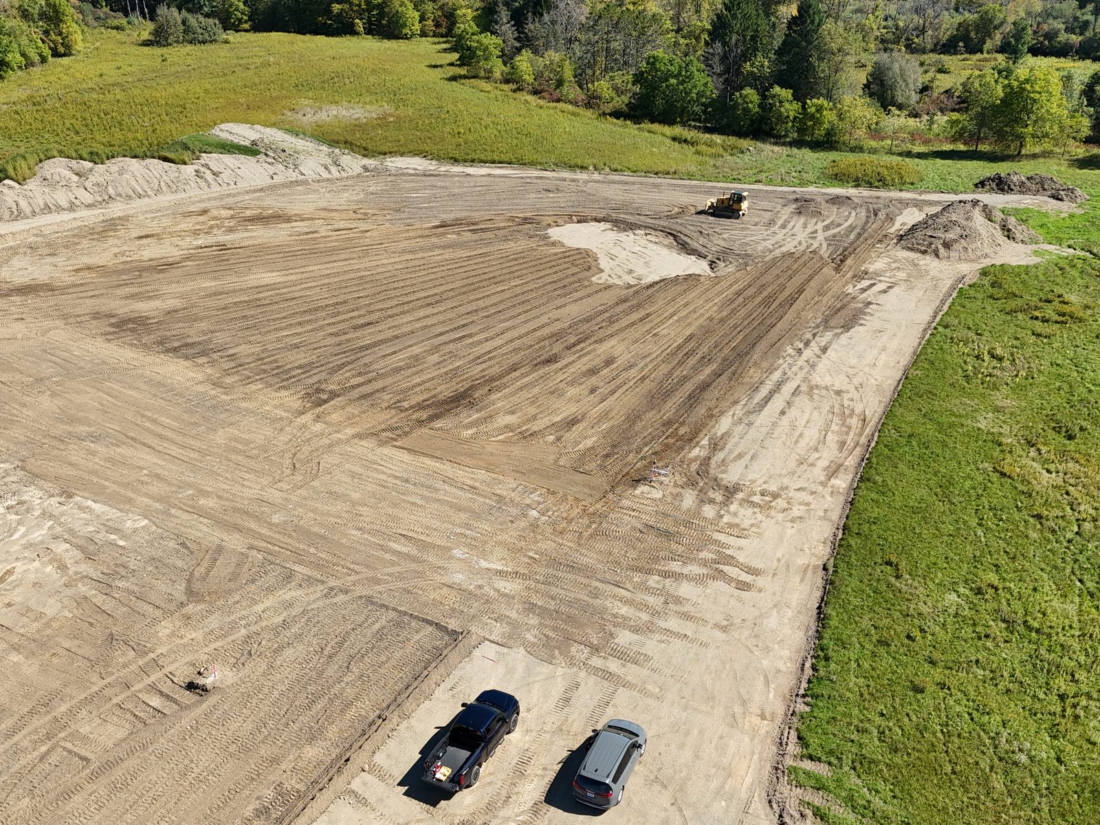 Aerial view of a dozer working a large graded area, with fresh track marks, two parked vehicles at the edge and meadow and trees around it