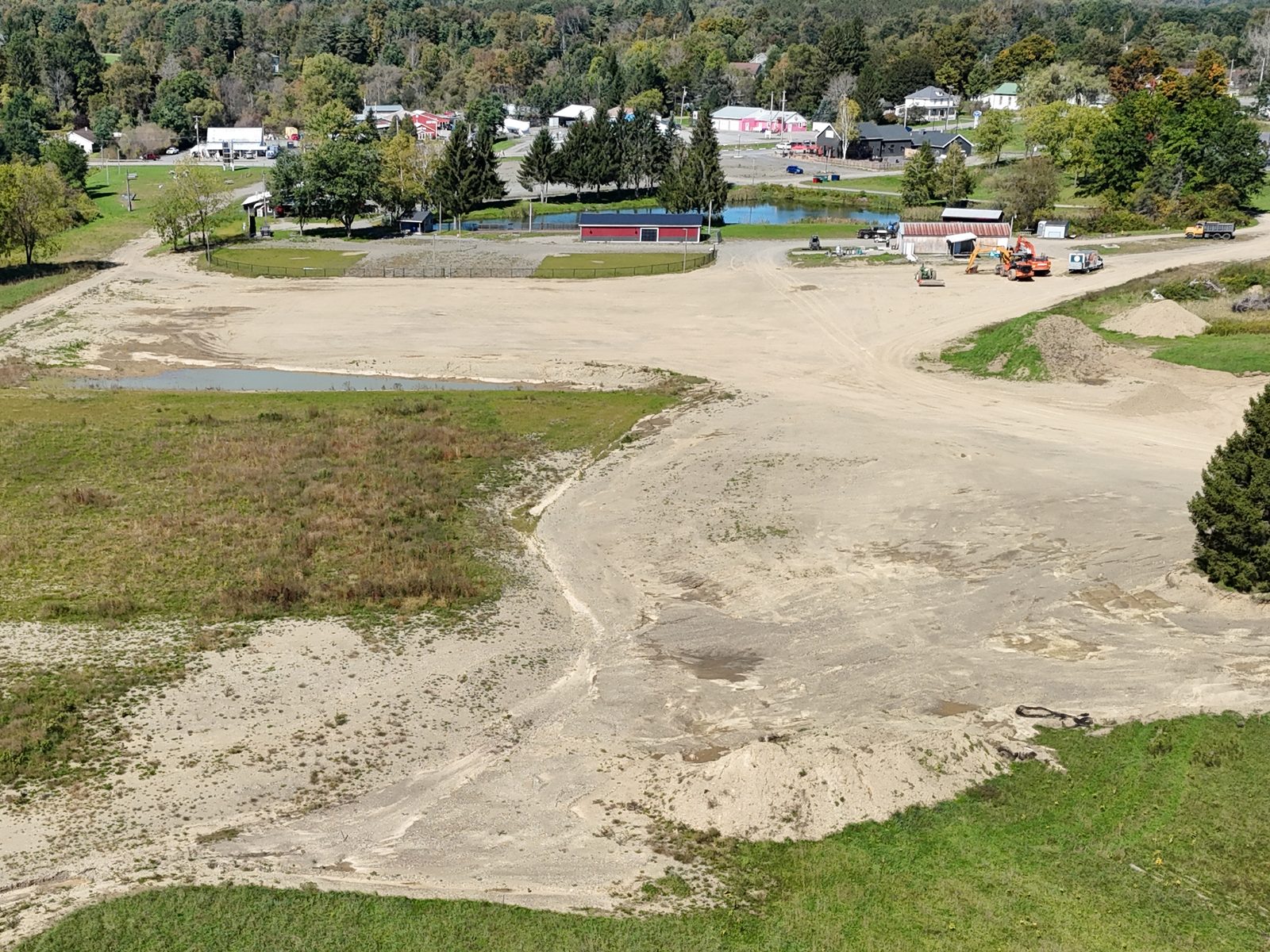 Aerial view across a large graded area toward the fenced wiffle ball fields, a red barn, the pond, construction equipment, and houses and businesses along the road beyond