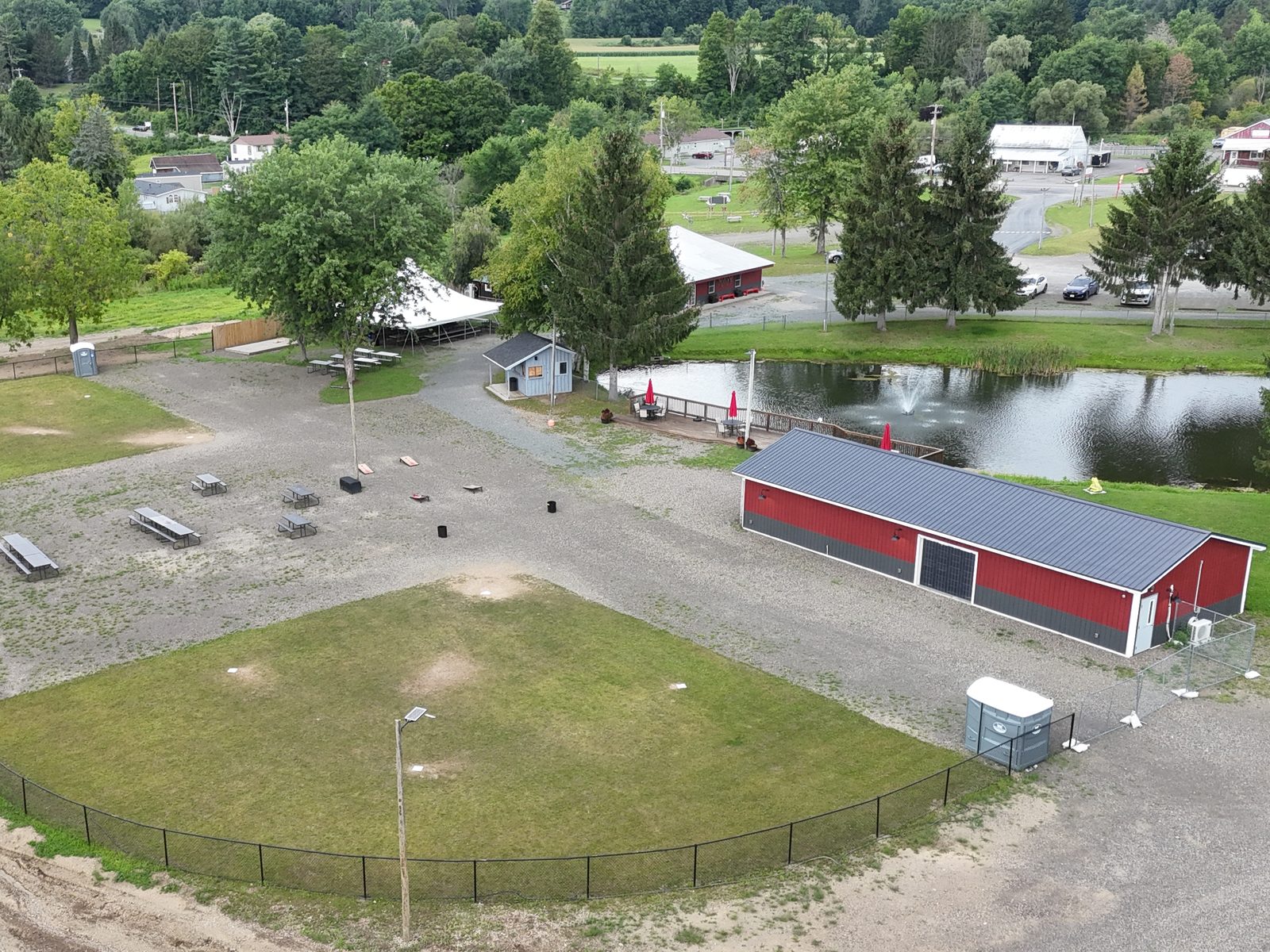Aerial view of a fenced wiffle ball field and picnic area, with a red barn, a pond with a fountain and tree-lined roads beyond