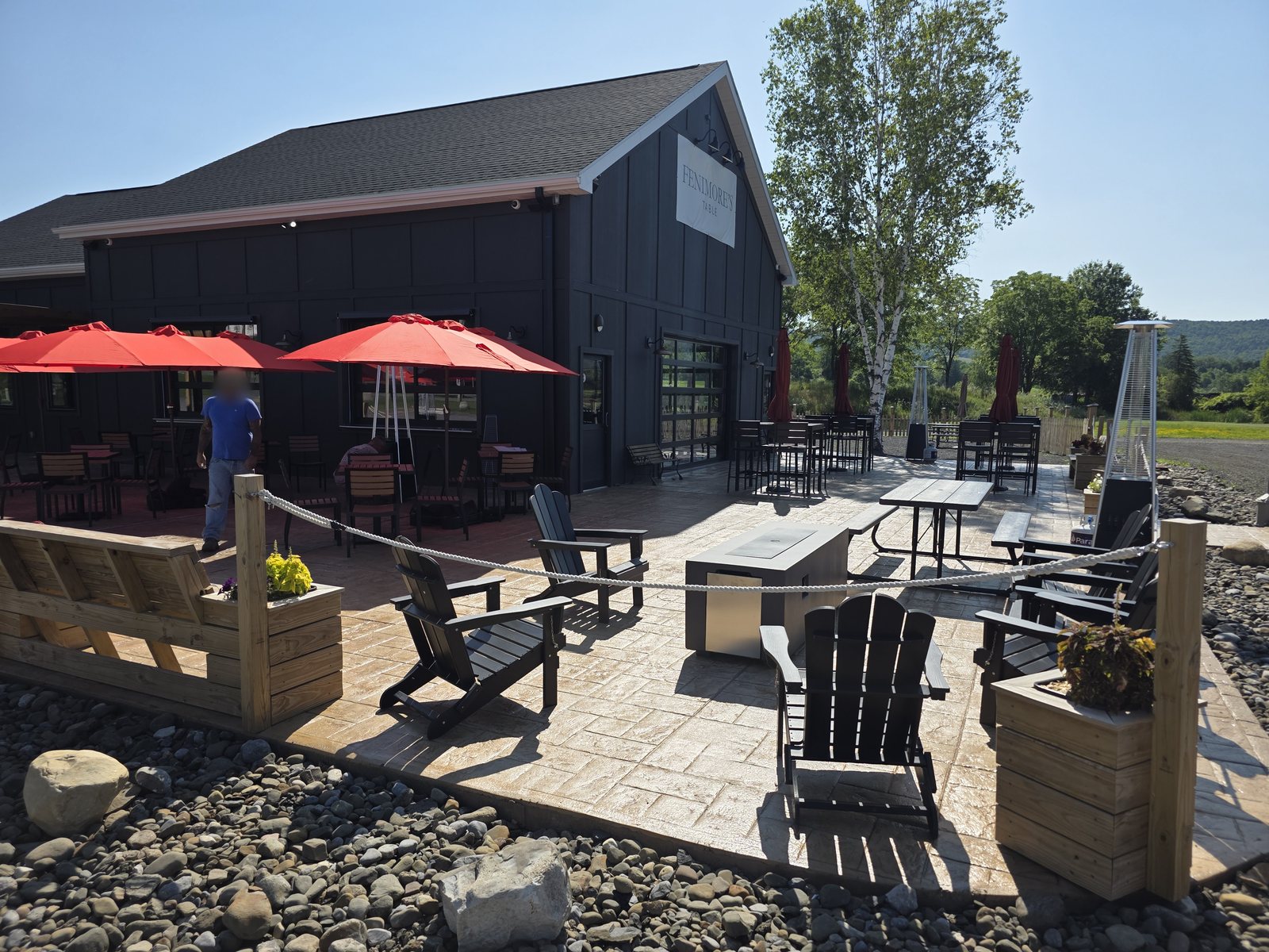 The Fenimore's Table patio on a sunny day, with Adirondack chairs around a fire table, red umbrellas and the Fenimore's Table sign on the building