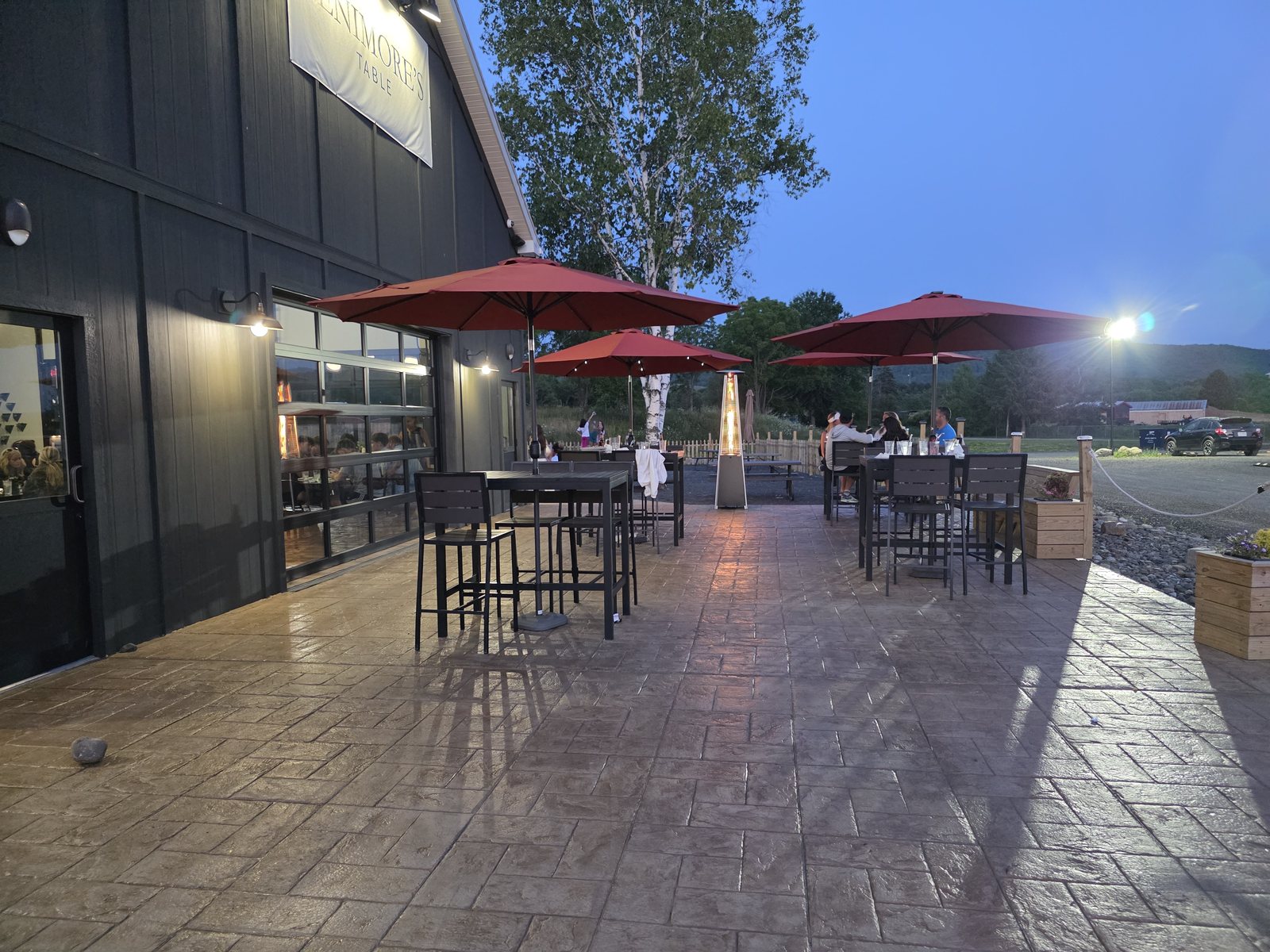 Guests at high-top tables on the Fenimore's Table patio at dusk, under red umbrellas beside the Fenimore's Table sign