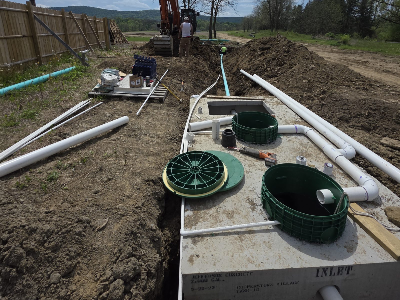 Concrete septic tank with green access risers and white pipe runs, excavator working the trench beyond