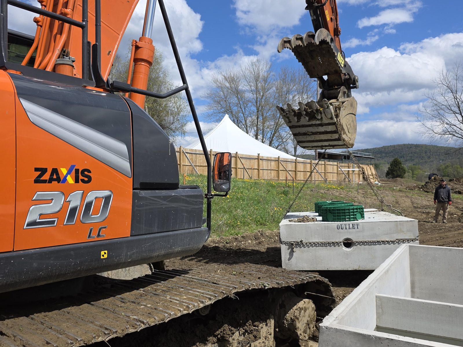 Hitachi excavator beside two precast concrete septic tanks, one marked Outlet